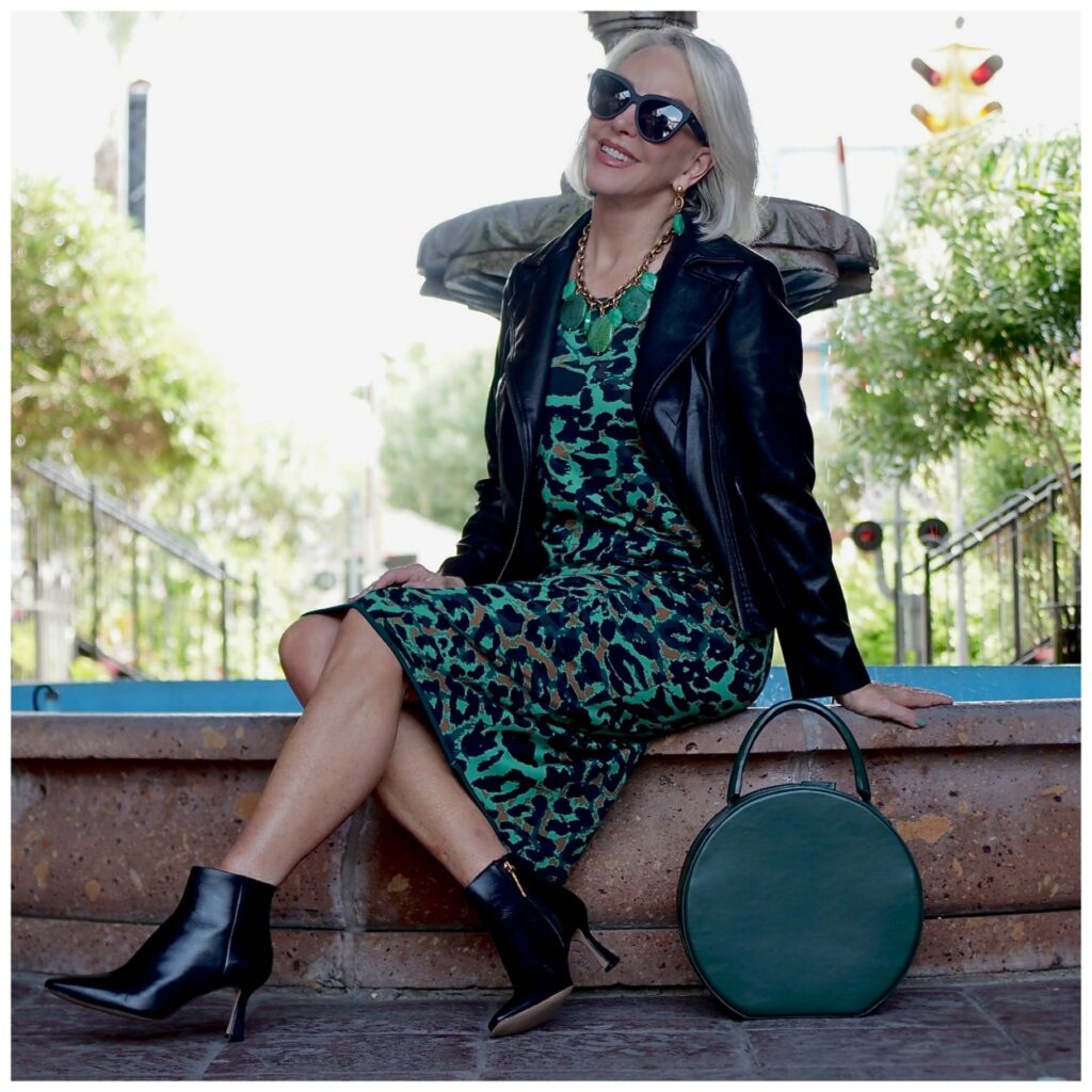 Sheree Frede of the SheShe Show sitting by fountain wearing a green leopard print dress with a black leather jacket.