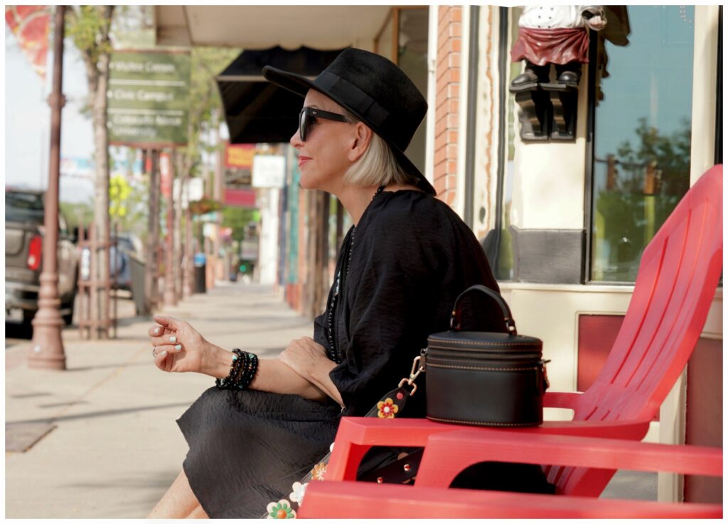 Sheree Frede of the SheSheShow sitting in a red adirondack chair on sidewalk front of a door wearing an