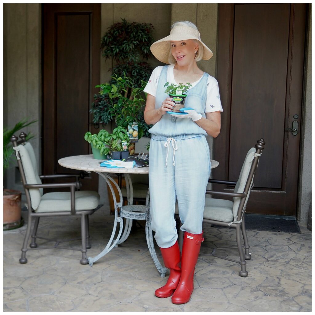 Sheree Frede of the SheShe Show standing by table holding a small plant. She is wearing a chambray jumpsuit and red hunter rain boots