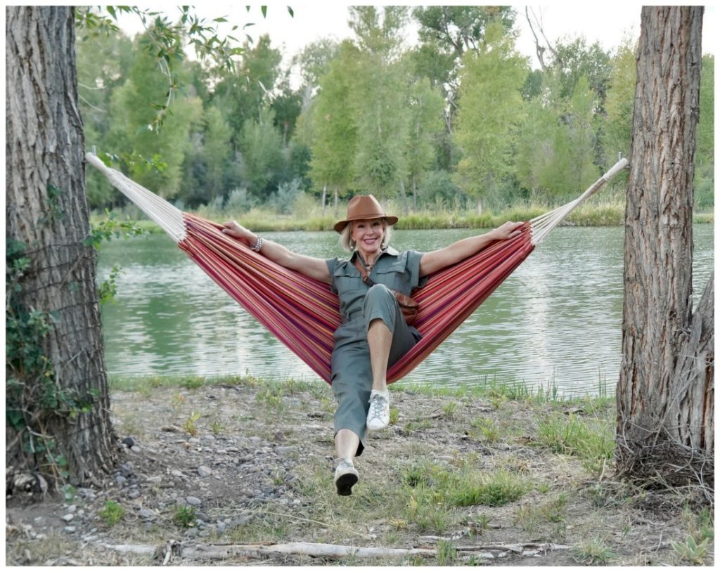 Sheree Frede of the SheShe Show lounging in a hammock by water wearing a brown leather hat and green utility jumpsuit