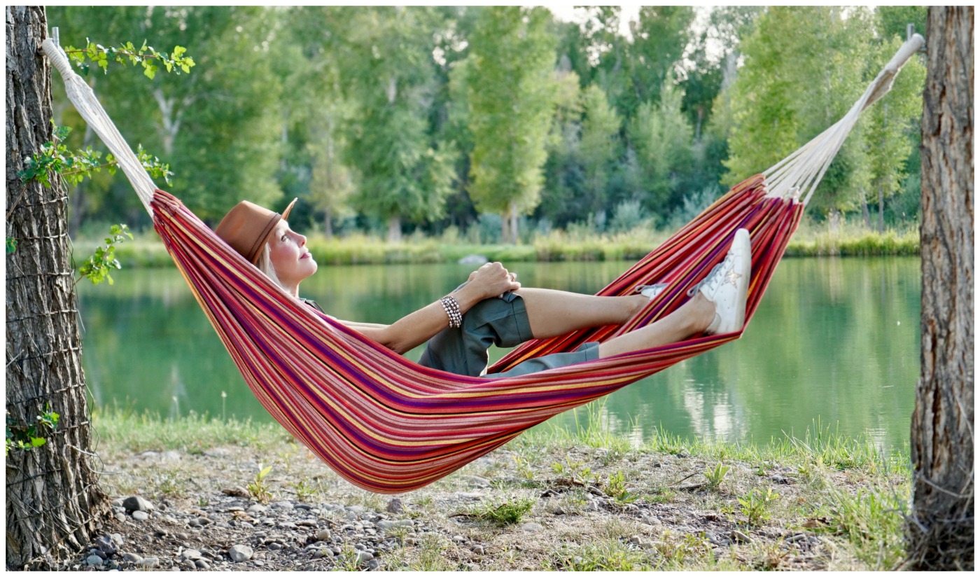 Sheree Frede of the SheShe Show lounging in a hammock by water wearing a brown leather hat and green utility jumpsuit