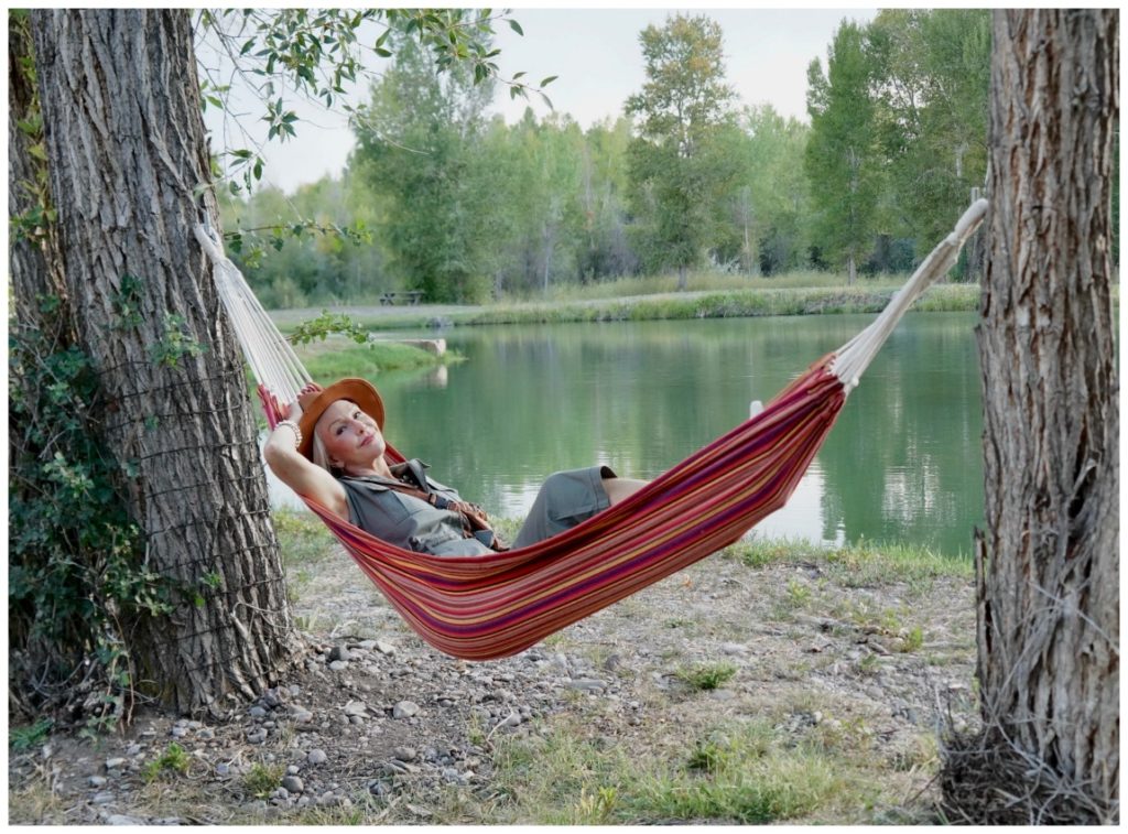 Sheree Frede of the SheShe Show lounging in a hammock by water wearing a brown leather hat and green utility jumpsuit