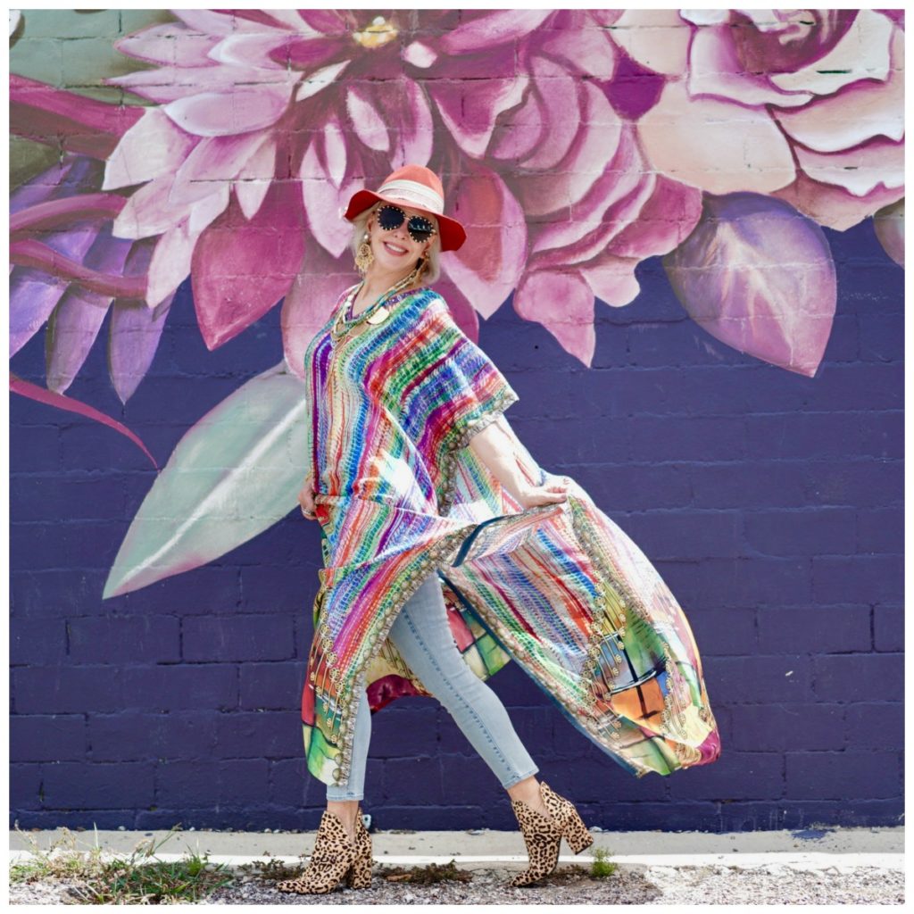 Sheree Frede of the SheShe Show walking in front of purple floral mural wearing a purple, green and orange print silk kaftan with brown leather hat