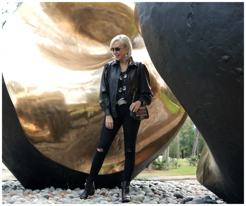 Sheree Frede of the SheShe Show standing in front of an outdoor sculpture wearing a black faux leather shirt and jeans