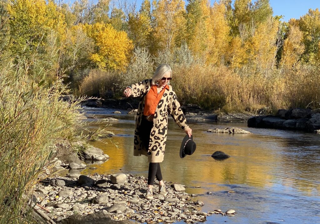 Sheree Frede of the SheShe Show standing on river rocks in the river wearing a lepard print cardigan, orange shirt and black jeans