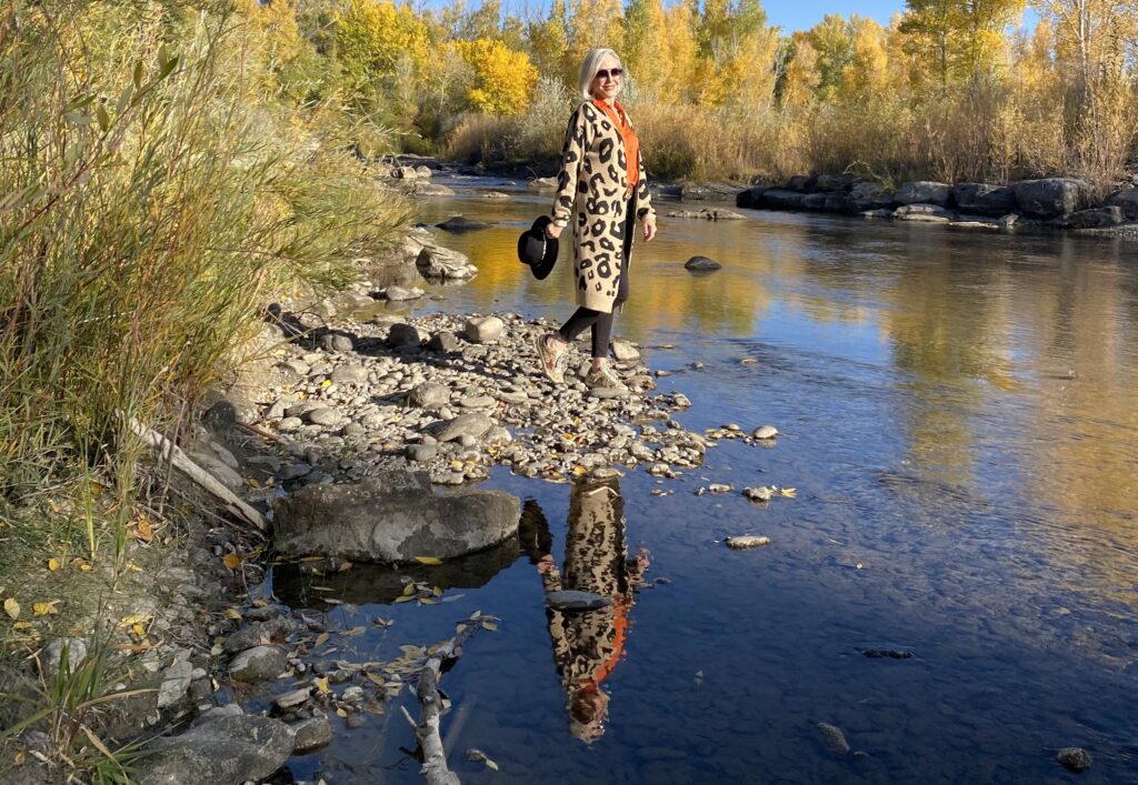 Sheree Frede of the SheShe Show standing on river rocks in the river wearing a lepard print cardigan, orange shirt and black jeans