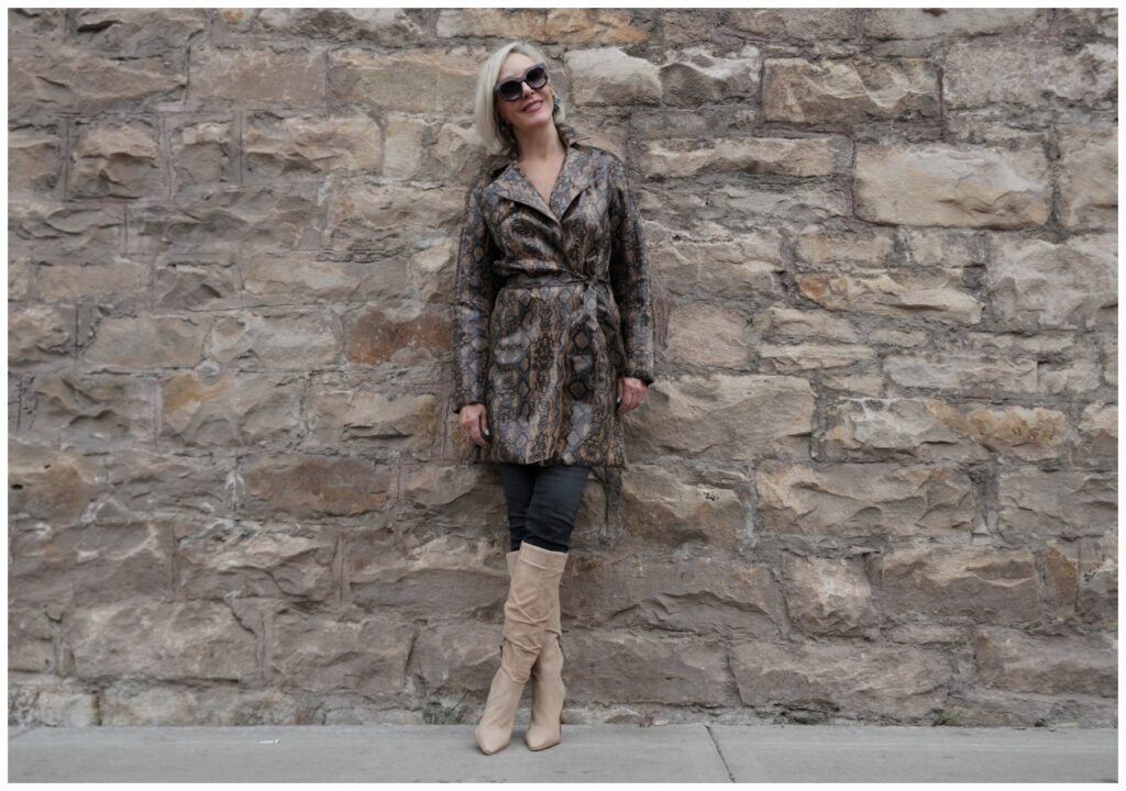 Sheree Frede of the SheShe Show standing in front of rock wall wearing a snakeprint trench coat with tan knee boots