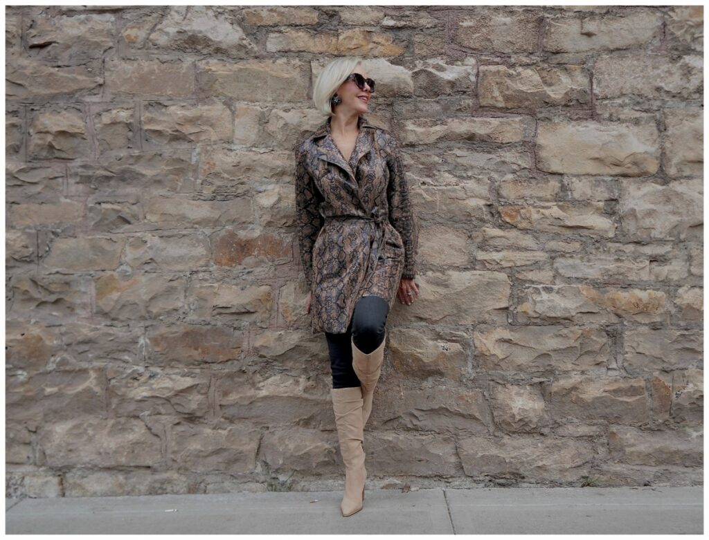 Sheree Frede of the SheShe Show standing in front of rock wall wearing a snakeprint trench coat with tan knee boots