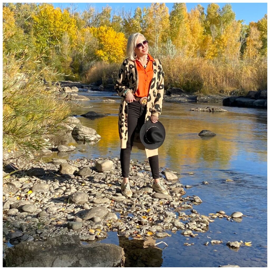 Sheree Frede of the SheShe Show standing on river rocks in the river wearing a lepard print cardigan, orange shirt and black jeans