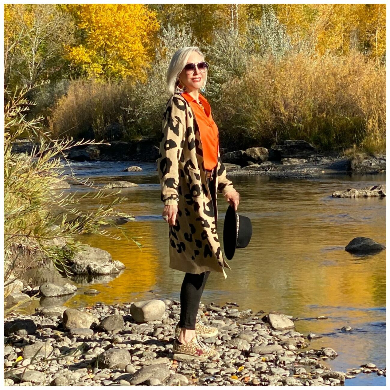 Sheree Frede of the SheShe Show standing on river rocks in the river wearing a lepard print cardigan, orange shirt and black jeans