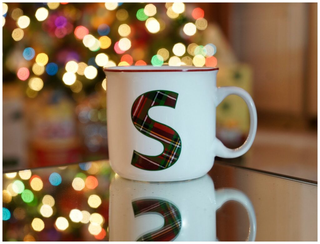 White coffee cup with a large tartan print S on the front with Chritsmas tree in the background.