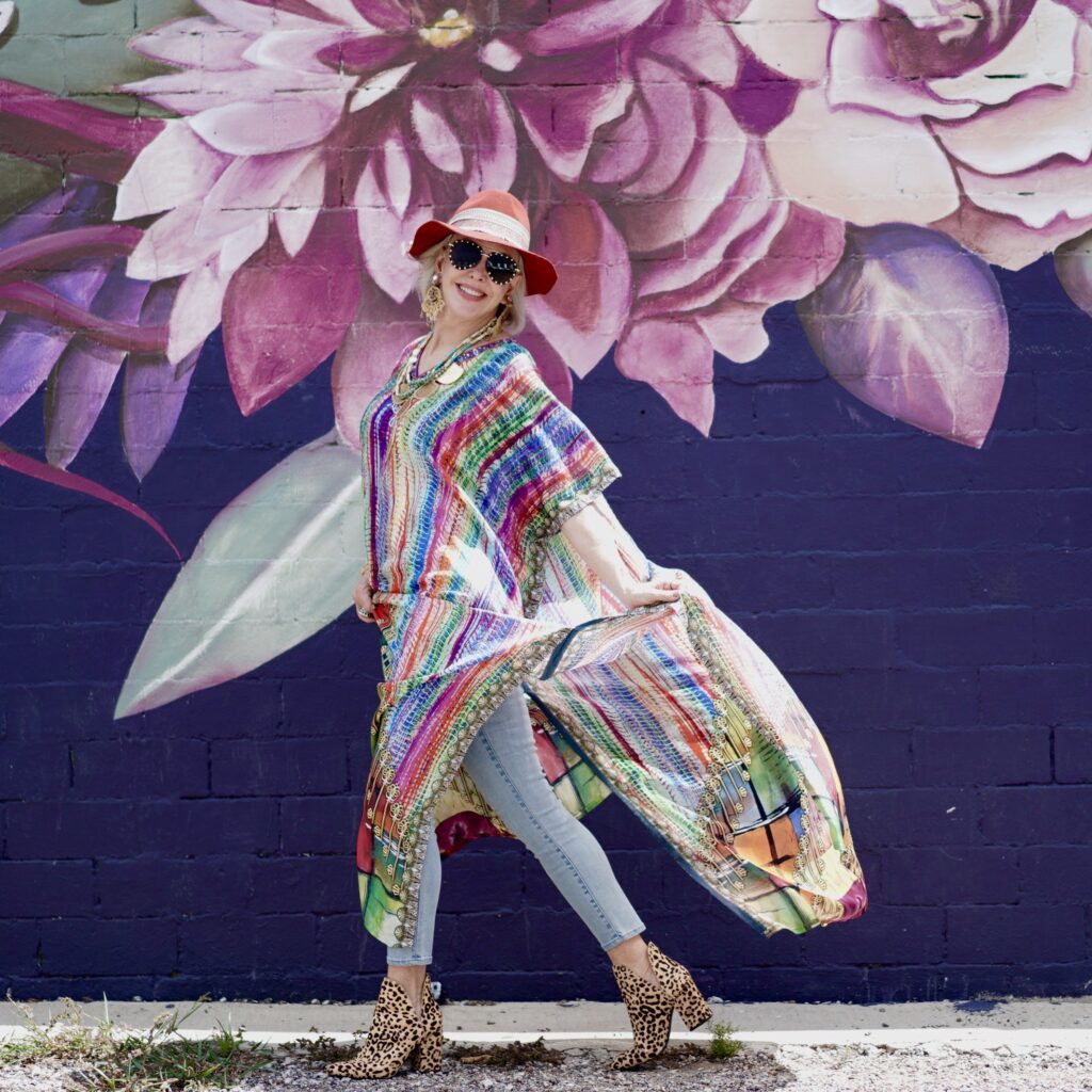 Sheree Frede of the SheShe Show walking in front of floral art wall wearing a multi colored strip kaftan and hat