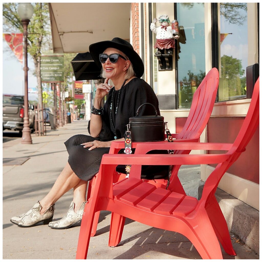 Sheree Frede of the SheShe SHow wearing a black dress sitting in a read chair on a sidewalk