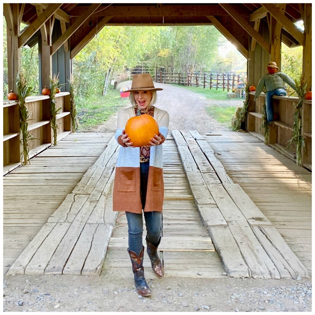 Sheree Frede of the SheShe Show standing on covered bridge holding a pumkin, wearing a long cardigan, Jeans and hat