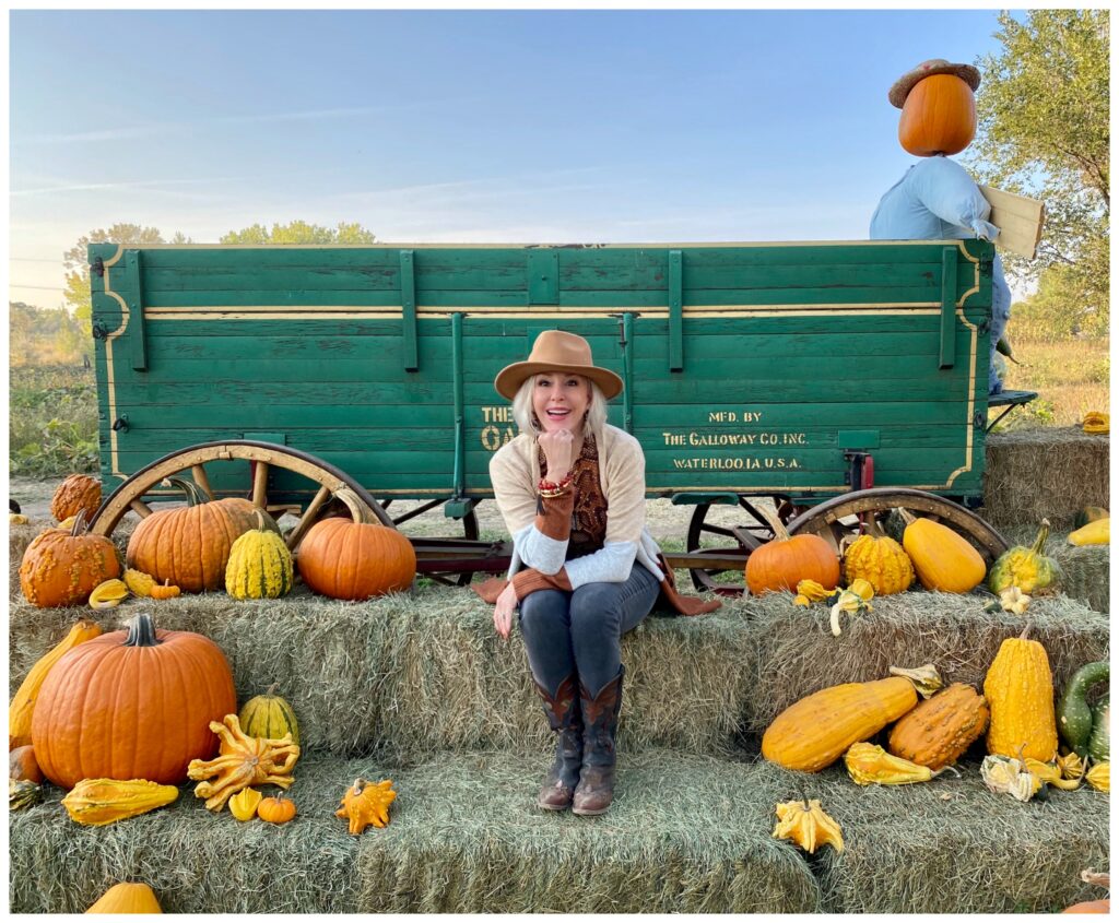 Sheree Frede of the SheShe Show sitting on a bale of hay by an old wagon wearing jeans, boots, hat and long cardigan