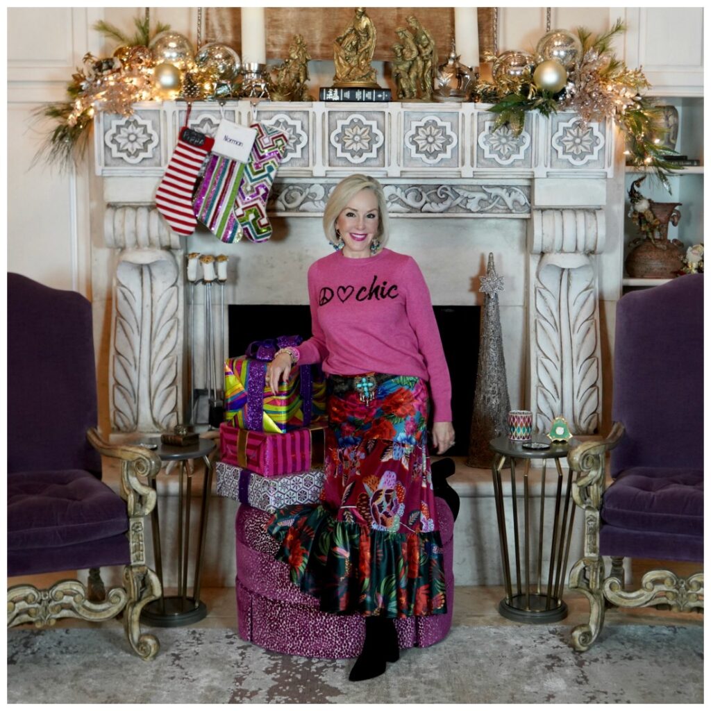 Sheree Frede of the SheShe Show standing in front of fireplace wearing a bright pink sweater and floral skirt