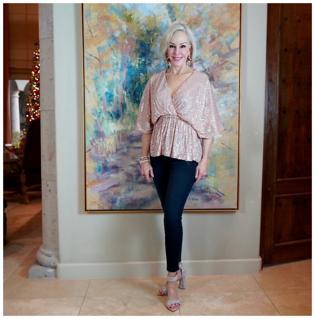 Sheree Frede of the SheShe Show standing in front of large art piece wearing a blush colored sequin top and blue jeans