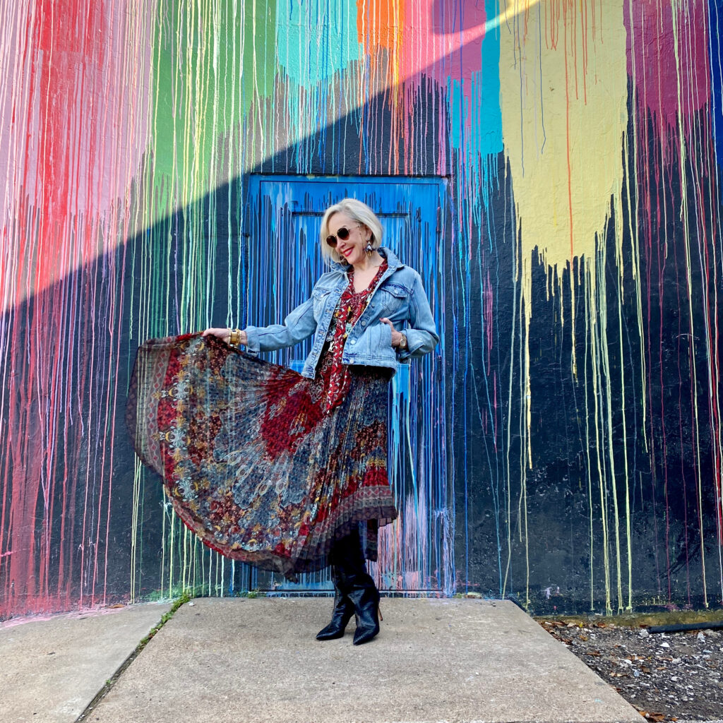 sheree Frede of the SheShe Show standing in front of paint drip mural wall wearing a flowy print skirt, knee boots and denim jacket