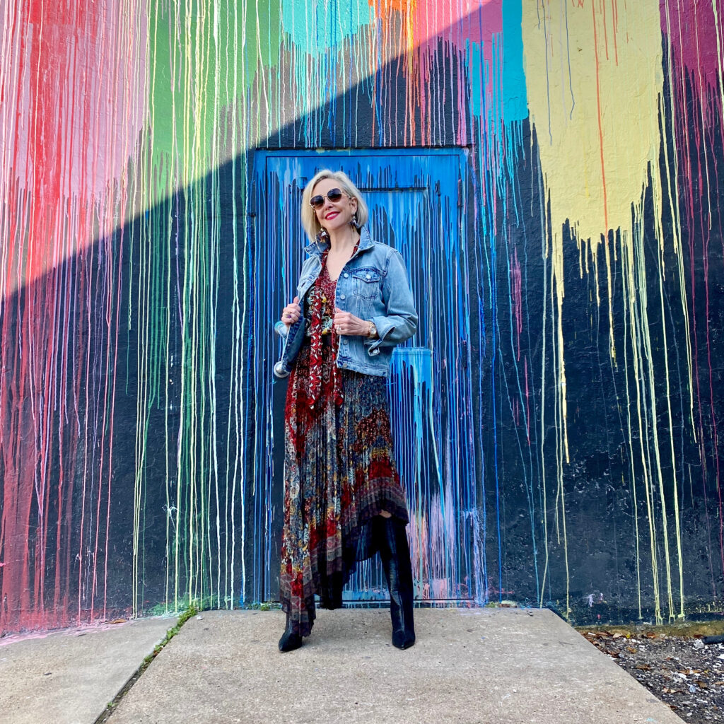 sheree Frede of the SheShe Show standing in front of paint drip mural wall wearing a flowy print skirt, knee boots and denim jacket