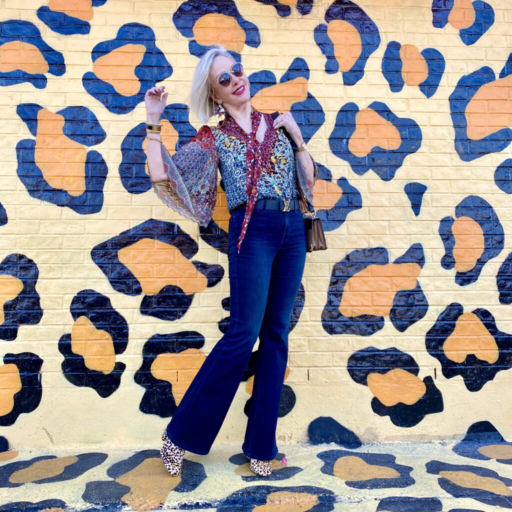 sheree Frede of the SheShe Show standing in front of paint drip mural wall wearing a flowy print bow blouse with flared jeans