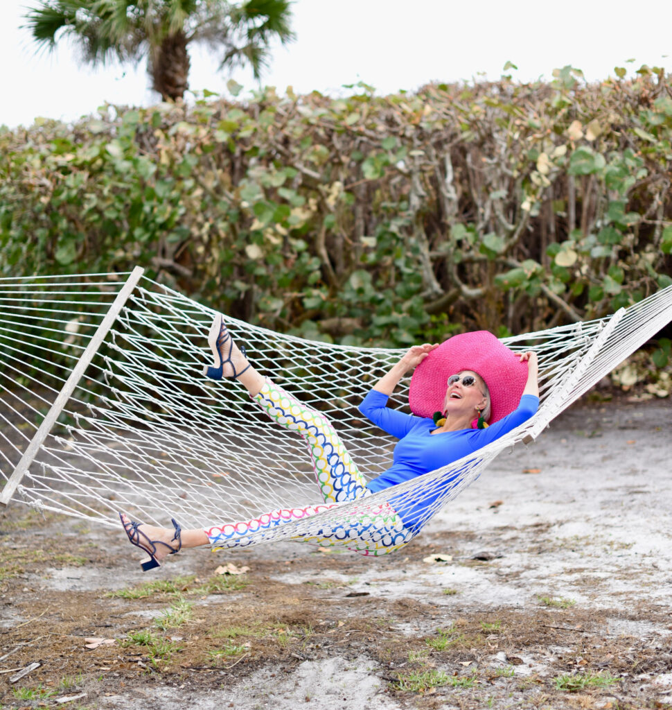 Sheree Frede of the SheSheShow, swinging in hammock blue longsleeve top, white pants with multi-colored pattern, large hot pink hat, denim blue sandals