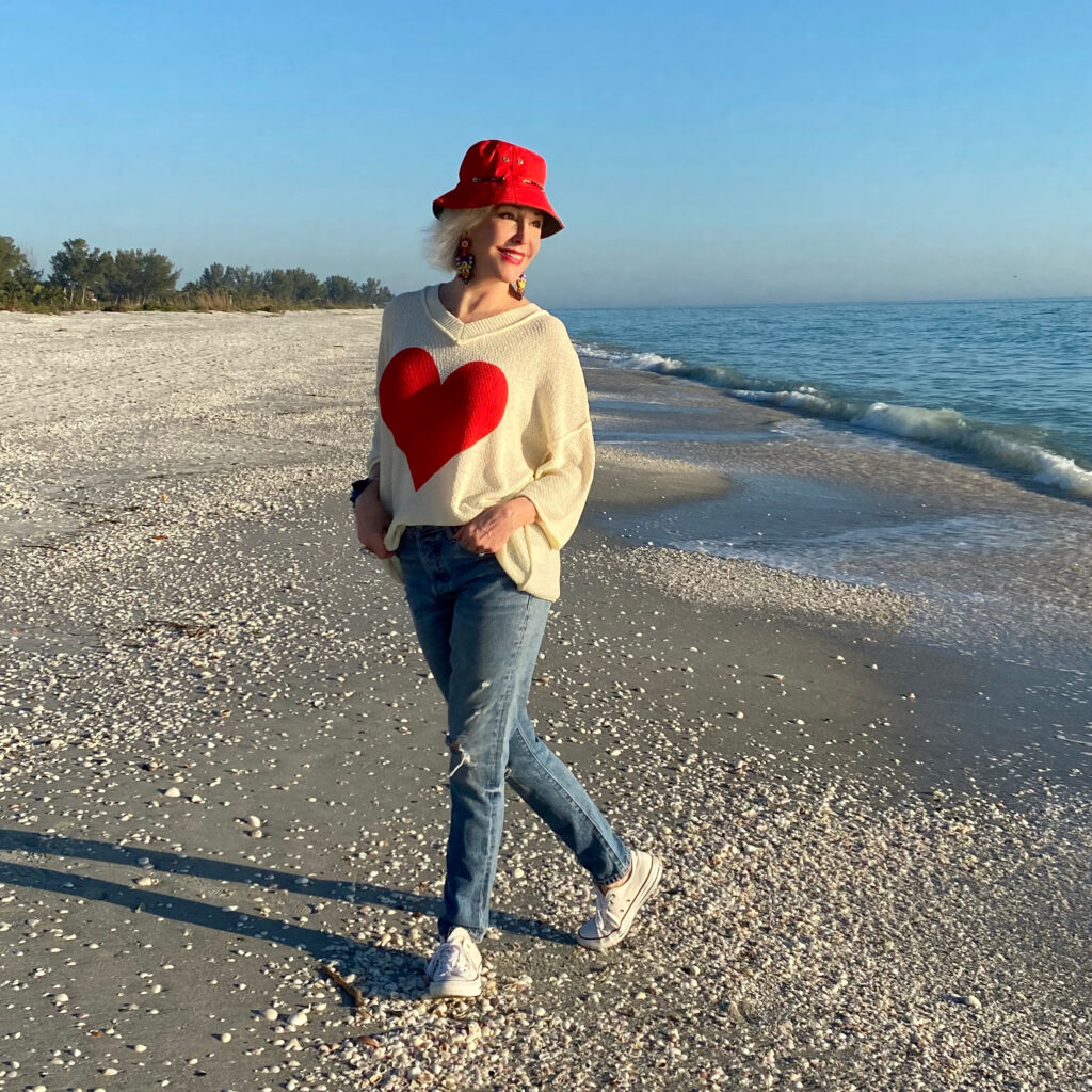 Sheree Frede of the SheShe Show walking on the brach wearing a cream sweater with a big red heart on the front with jeans and sneakers