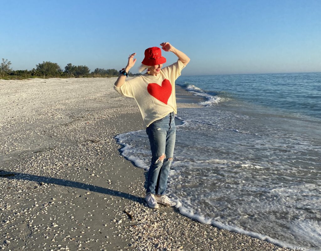 Sheree Frede of the SheShe Show walking on the brach wearing a cream sweater with a big red heart on the front with jeans and sneakers