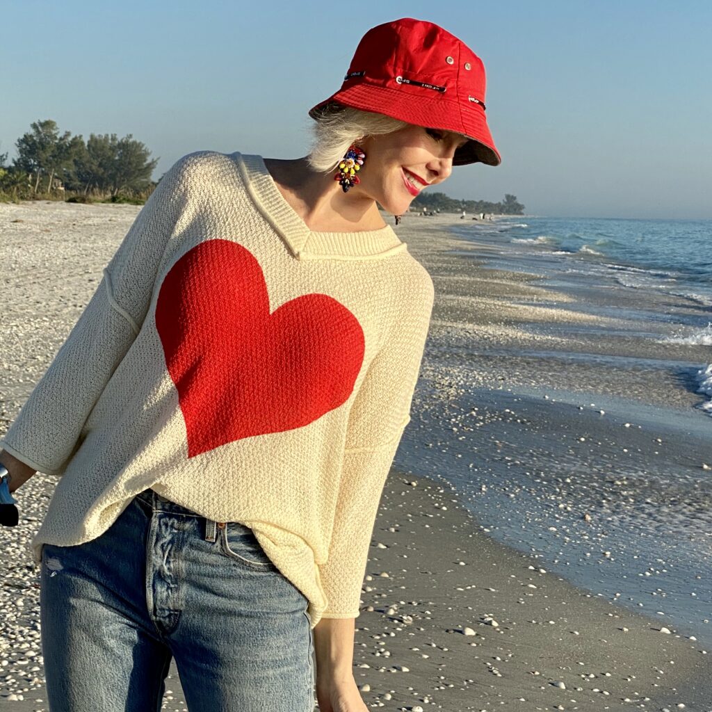 Sheree Frede of the SheShe Show walking on the brach wearing a cream sweater with a big red heart on the front with jeans and sneakers