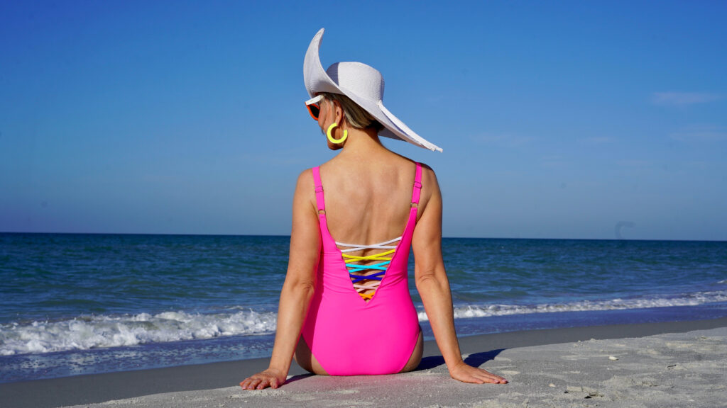 Sheree Frede of the SheShe Show in the water on the beach wearing a big hat and hot pink swimsuit