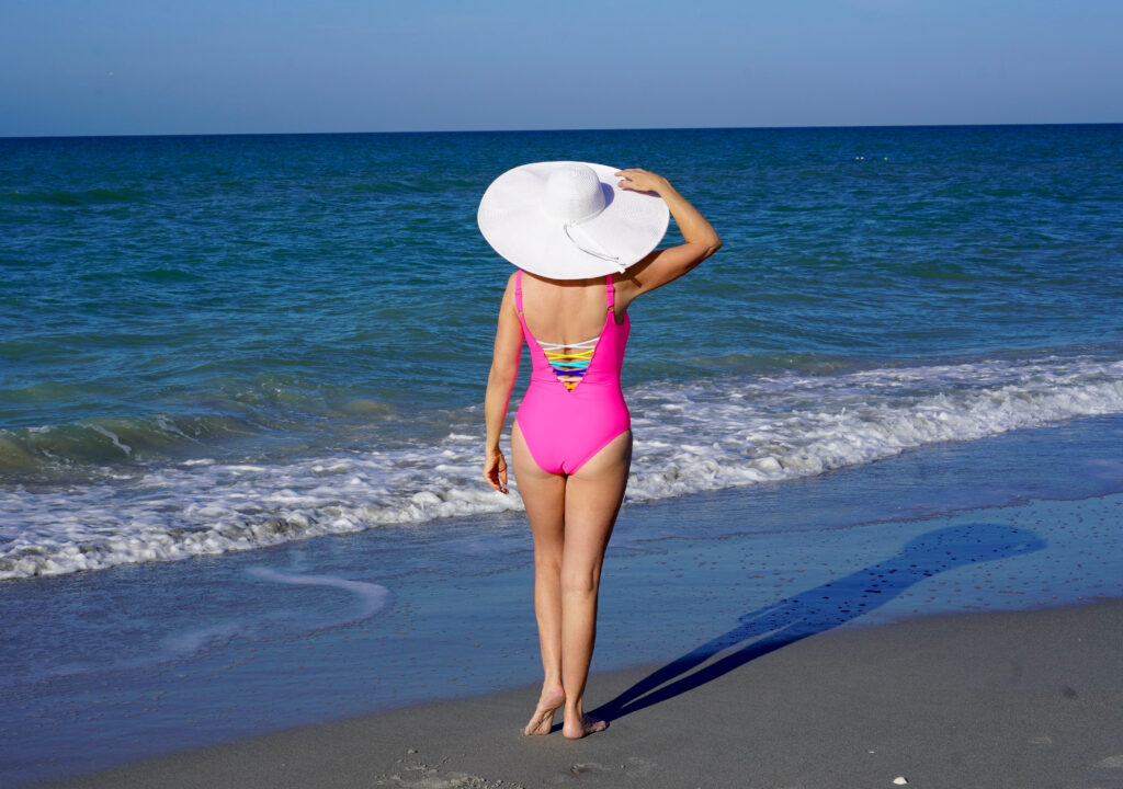 Sheree Frede of the SheShe Show in the water on the beach wearing a big hat and hot pink swimsuit