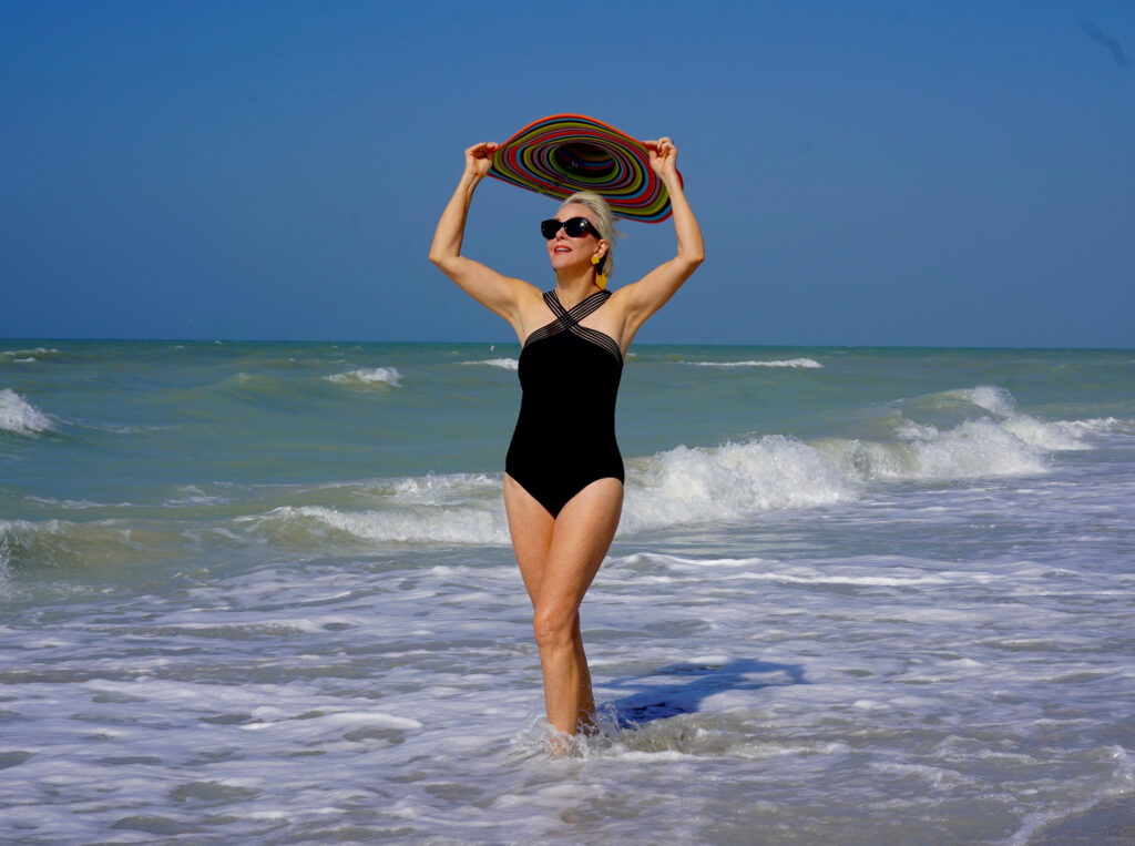 Sheree Frede of the SheShe Show in the water on the beach wearing a big hat and black one piece swimsuit