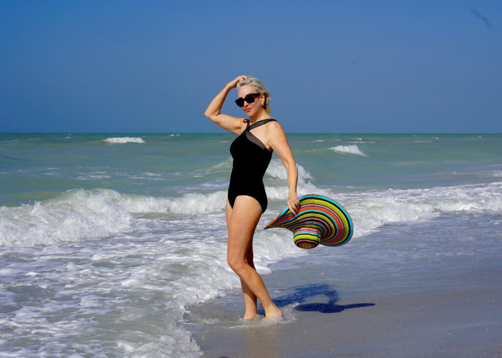 Sheree Frede of the SheShe Show in the water on the beach wearing a big hat and black one piece swimsuit