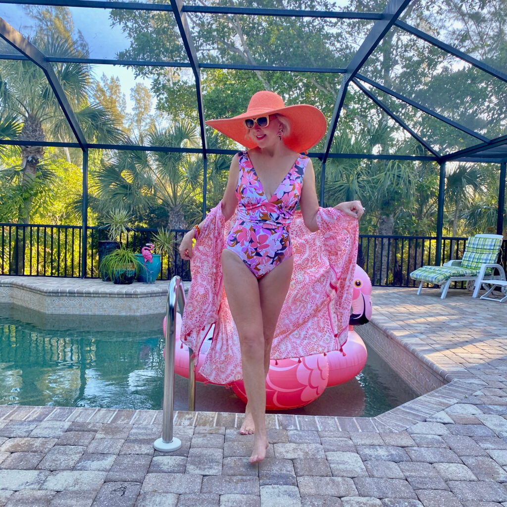Sheree Frede of the SheShe Show in the water on the beach wearing a big hat and pink floral one piece swimsuit