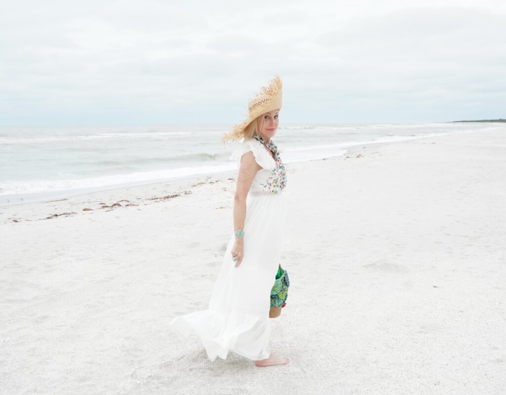 Sheree Frede of tSheree Frede of the SheShe Show walking on the beach weraring a long white dress and straw hat