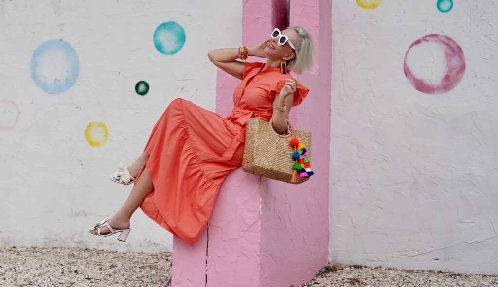 Sheree Frede of the SheShe Show stinnon on a pink and white wall wearing a bright orange maxi dress