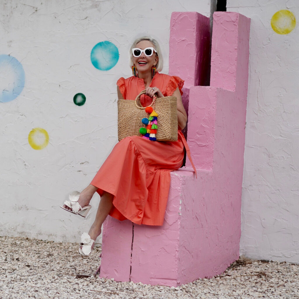 Sheree Frede of the SheShe Show stinnon on a pink and white wall wearing a bright orange maxi dress