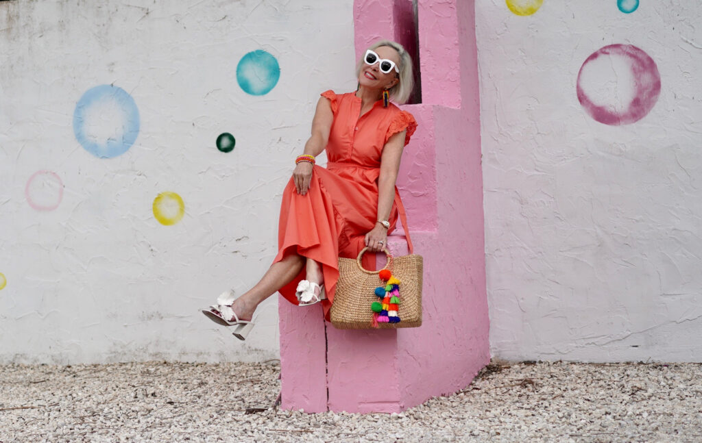 Sheree Frede of the SheShe Show stinnon on a pink and white wall wearing a bright orange maxi dress