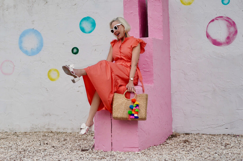 Sheree Frede of the SheShe Show stinnon on a pink and white wall wearing a bright orange maxi dress