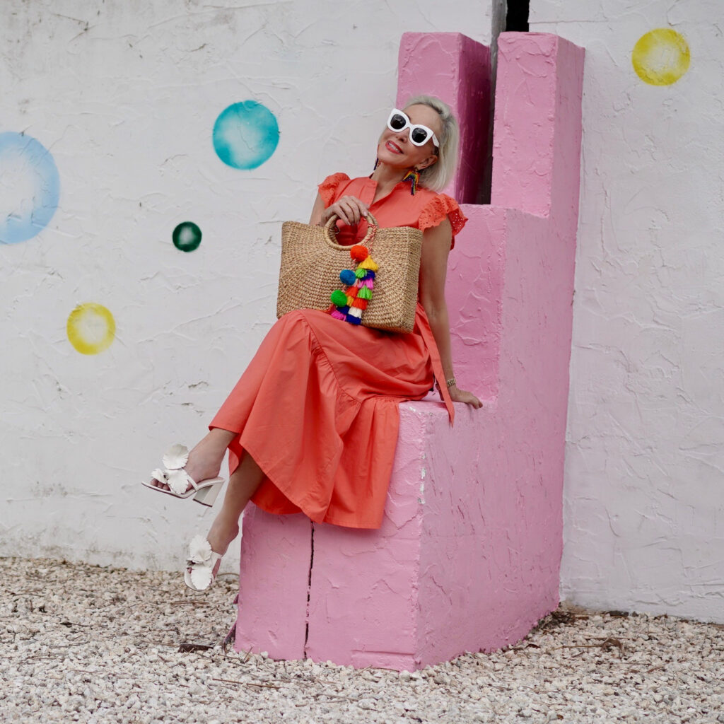 Sheree Frede of the SheShe Show stinnon on a pink and white wall wearing a bright orange maxi dress