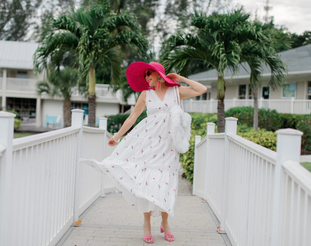 Sheree Frede of the SheShe Show walking on white bridge wearing a white maxi dress and hot pink hat