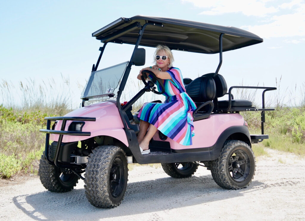 Sheree Frede of the SheShe Show wearing a multi color stripe midi dress with big white hat in pink golf cart