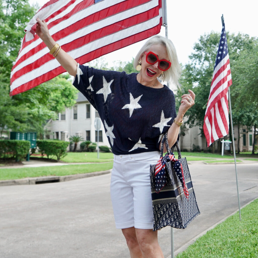 Sheree Frede of the SheShe Show standing by American Flags wearing white shorts and navy sweater with white stars