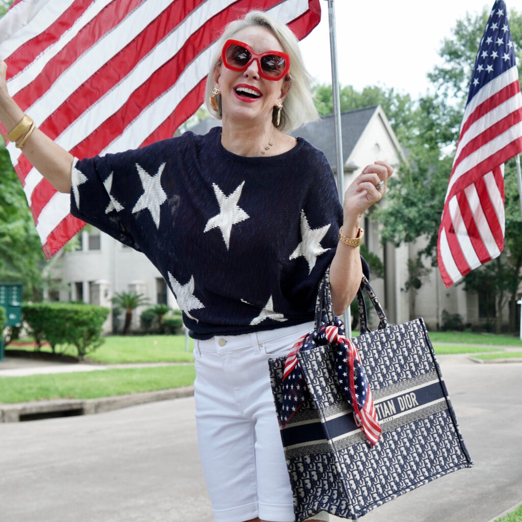 Sheree Frede of the SheShe Show standing by American Flags wearing white shorts and navy sweater with white stars