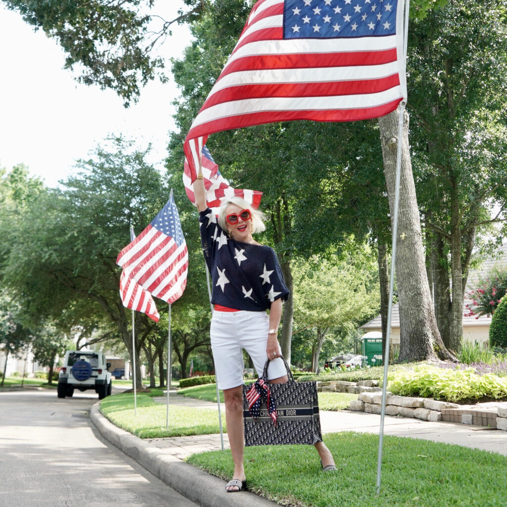Sheree Frede of the SheShe Show standing by American Flags wearing white shorts and navy sweater with white stars