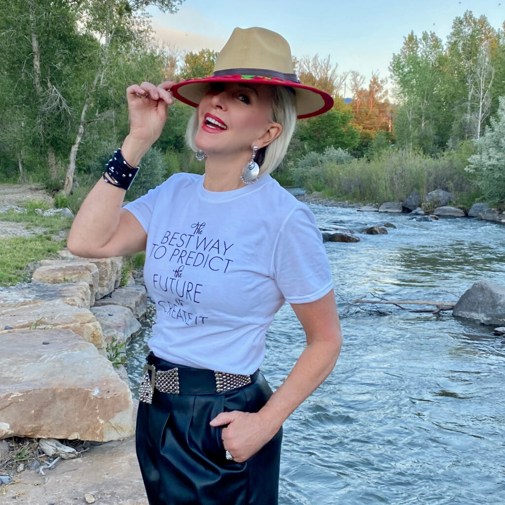 Sheree Frede of the SheShe Show by the river wearing a white graphic tee shirt with black faux leather shorts with a hat