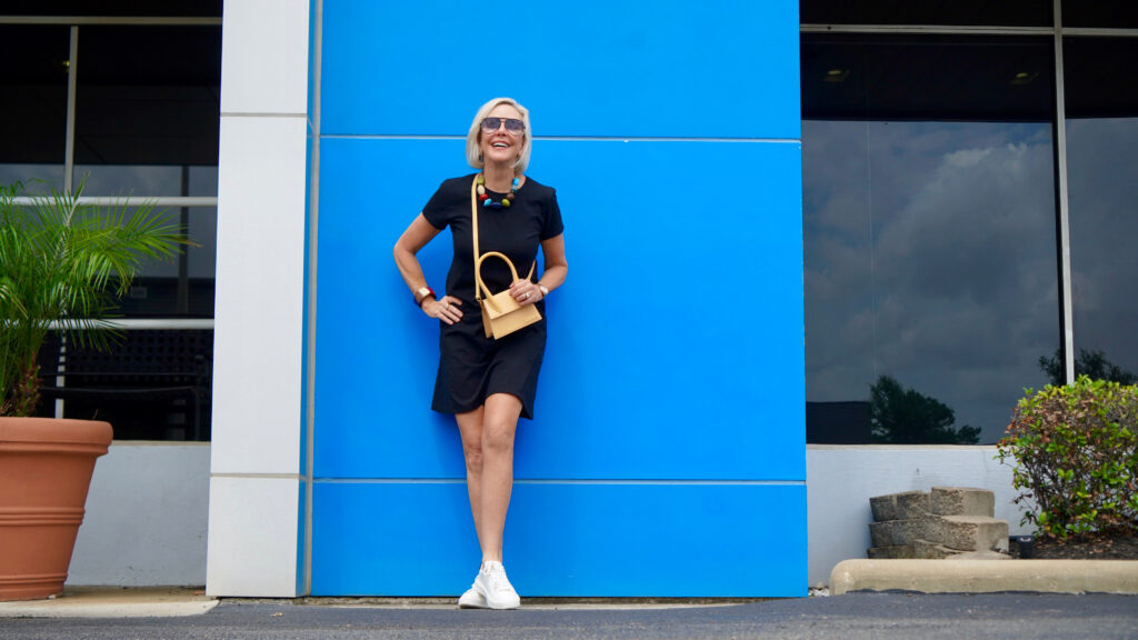 Sheree Frede of the SheSheShow standing in front of blue wall wearing a black short sleeve t-shirt dress