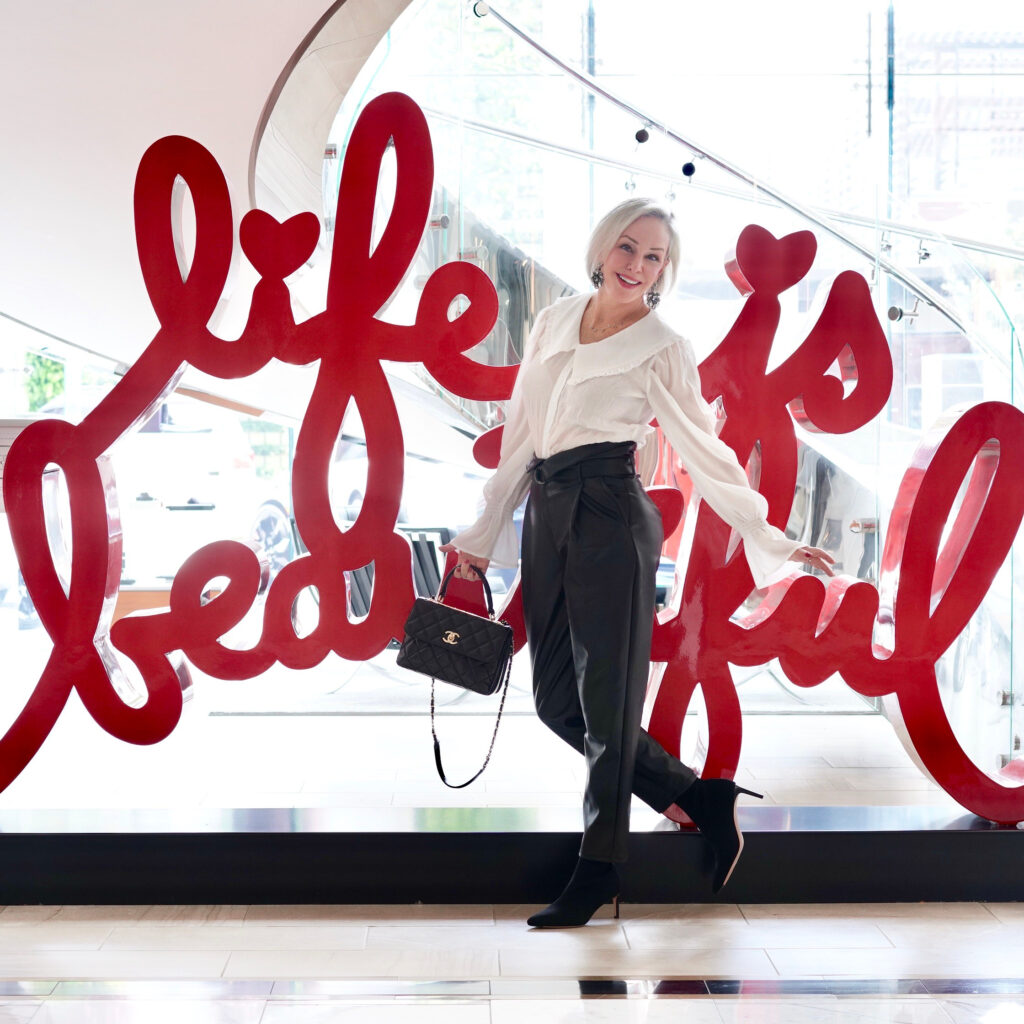 Sheree Frede of the SheShe Show standing in front of Life is Beautiful Sign wearing black faux leather joggers and feminine white top