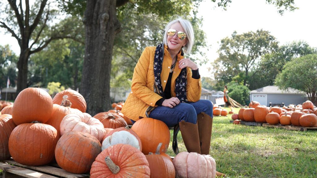 Sheree Frede of the SheShe Show sitting in a pumpkin patch wearing a yellow quilted barn jacket jeans and boots