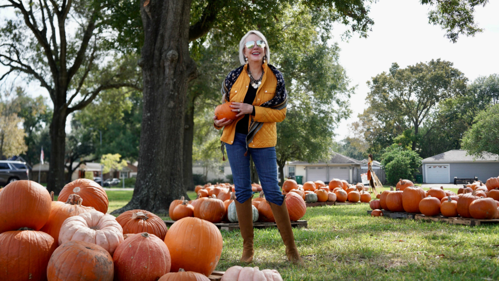 Sheree Frede of the SheShe Show sitting in a pumpkin patch wearing a yellow quilted barn jacket jeans and boots