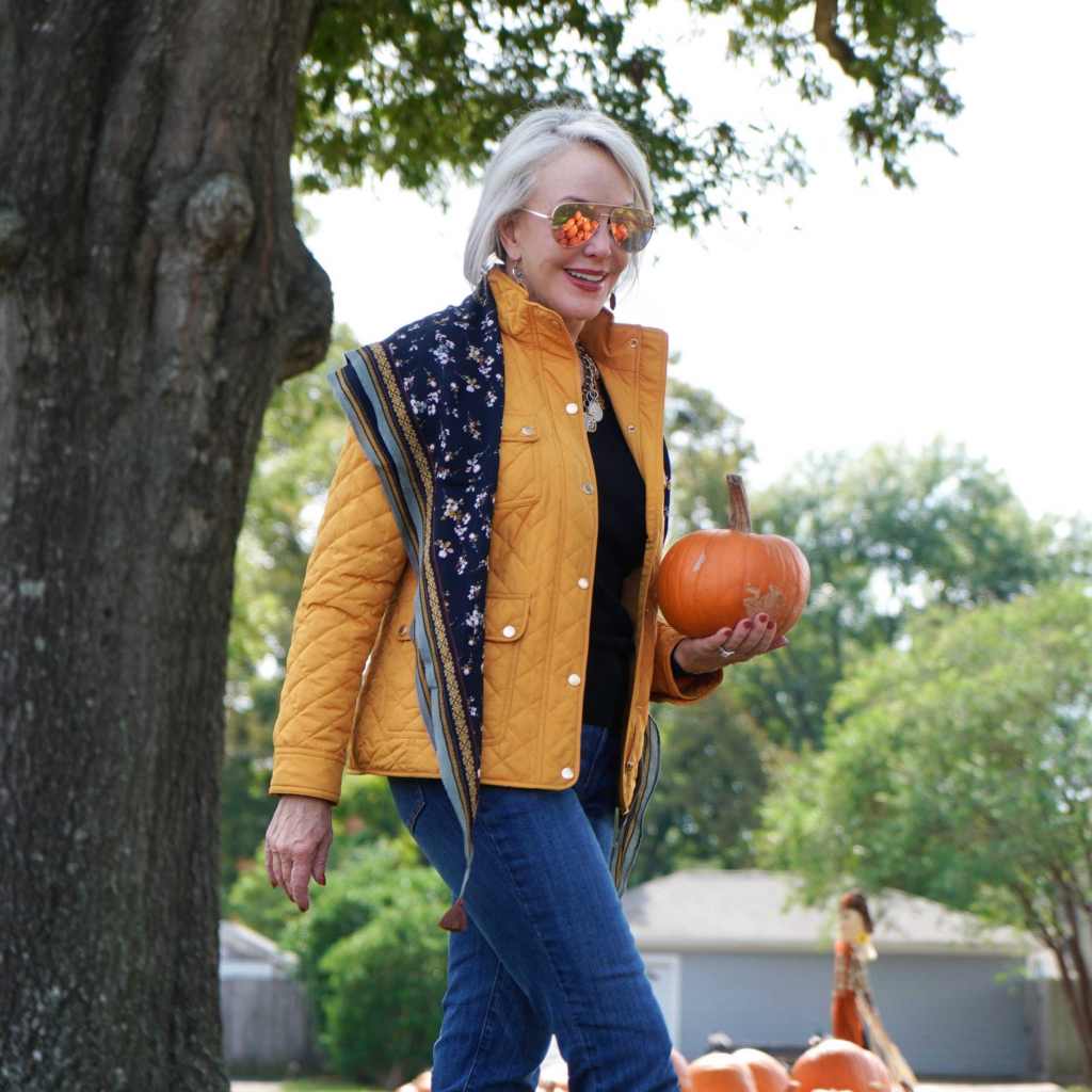 Sheree Frede of the SheShe Show sitting in a pumpkin patch wearing a yellow quilted barn jacket jeans and boots